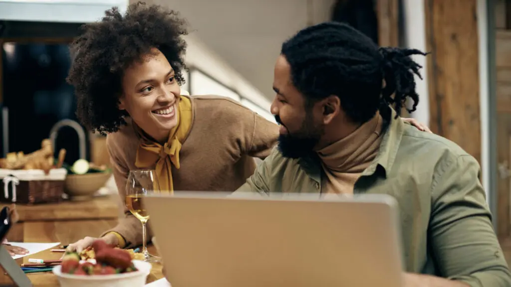 A woman and a man at the dinner table, laughing with their laptop.